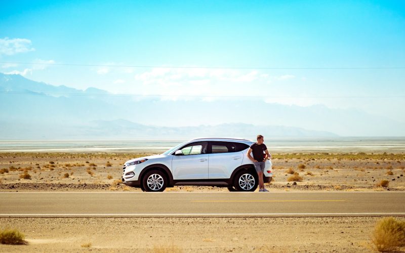 man standing beside white SUV near concrete road under blue sky at daytime