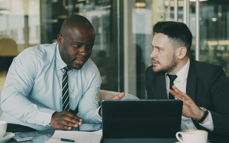 Two businessmen collaborating over a tablet and laptop